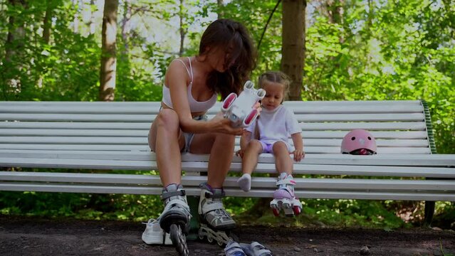 Mother and daughter taking off inline roller on bench after skating in public park together. Family leisure sport activity weekend.