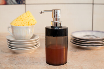 Composition about washing dishes. A bottle of dishwashing liquid against the background of plates and a cup with a sponge in the kitchen. An image about housework, kitchen care and cleanliness.