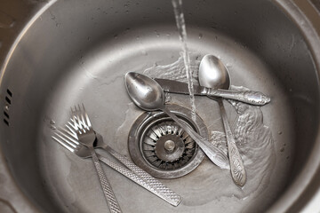 Composition about washing dishes. A jet of water from a tap pours into a washbasin with forks and spoons. An image about housework, kitchen care and cleanliness.