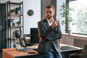 Leaning on the table. Businessman in suit is in the office with computer