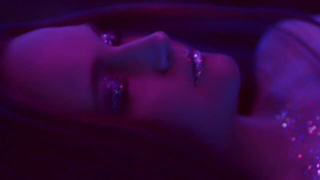 Vertical video. Neon girl. Glitter makeup. Nightclub beauty. Purple color light relaxed woman with sparkling face skin eyeshadow lips dancing with closed eyes on dark.