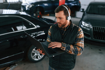 Standing with laptop in hands and in headphones. Man in uniform is working in the auto salon
