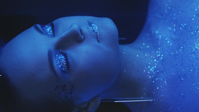 Vertical video. Sparkling makeup. Neon beauty. Disco look. Night portrait of blue color light woman face with shiny glitter skin eyeshadow lips on dark.