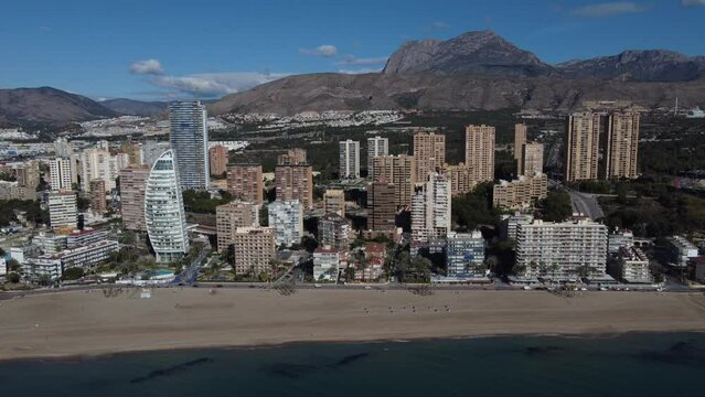 Sideways aerial reveal of the Poniente beach in Benidorm. Puigcampana and Aitana mountains in the background.