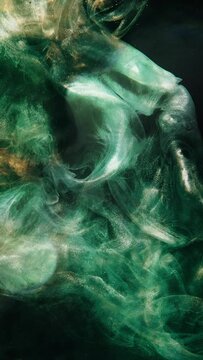 Vertical video. Ink water. Glitter fluid. Underwater splash. Abyss void. Green gold color glowing shiny dust particles smoke cloud floating on dark black abstract background.