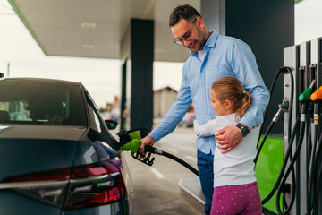 The father pours fuel into the car while his daughter hugs him and keeps him company