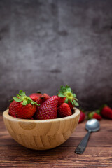 Sweet red strawberries in a wooden bowl placed on a table. Teaspoon, vertical image with selective focus.