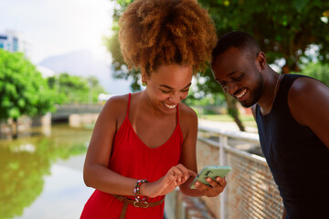 young afro black brazilian heterosexual couple using a cell phone and having fun outdoors in a park...