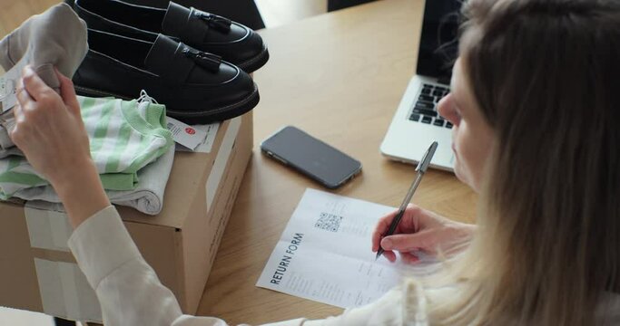 A young woman writes a return order and sits at a table with a laptop at home. Close-up of a female customer writing on paper and filling out a form, wanting to return a goods.