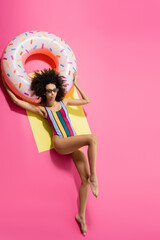 top view of barefoot african american woman in swimwear and stylish sunglasses getting tan near inflatable ring on yellow and pink.