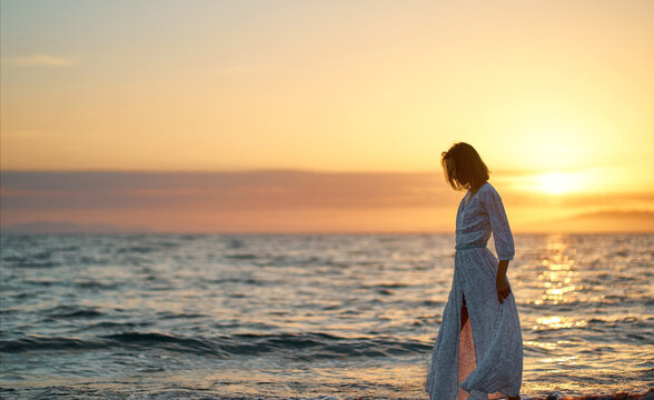 Beautiful young woman in long elegant dress at sunset. Silhouette of dreamy girl walking along the sea