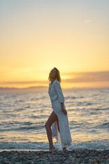 Attractive young elegant lady in dress enjoying sunset time at the beach. Background orange sundown on the sea