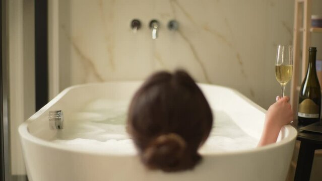 Back view of unrecognizable young woman with dark hair lying in luxury white bathroom holding glass of champagne. Closeup tracking shot of gorgeous female relaxing in bath. Shooting in slow motion.