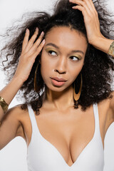 young african american model adjusting curly hair while looking away isolated on grey.