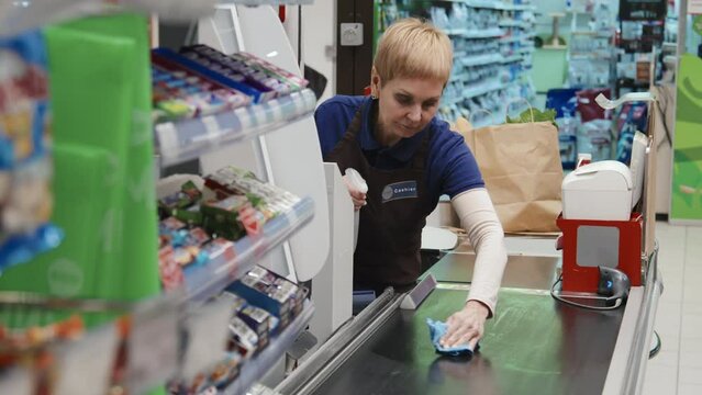 Mature cashier in uniform cleaning her workplace with rag standing behind the counter in supermarket