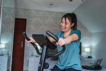 Fototapeta premium A young woman riding indoor bike during the day and drinking water and wiping with towel