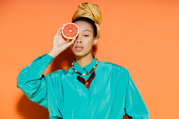 Fashionable african american woman with headscarf holding grapefruit on orange background.