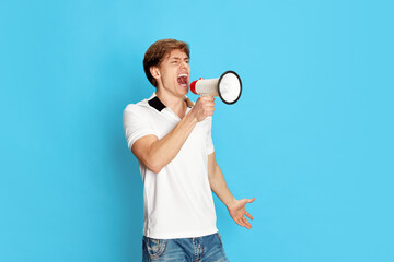 Portrait of young man, activist, emotionally shouting in megaphone against blue studio background....