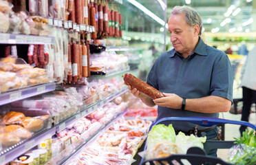 Elderly male customer looking for sausage in meat section of supermarket