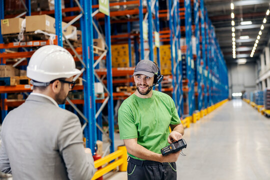 A warehouse man is holding bar code reader and showing loaded goods to his superior.
