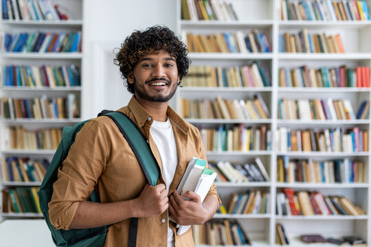 Portrait of happy satisfied african american student with curly hair, guy with books and backpack standing in university academic library smiling and looking at camera.