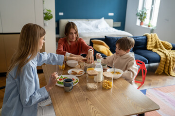 Single mom with children eating in morning. Pleased kid boy holding spoon cocoa while mom and teen sister want drink coffee. Family varied healthy breakfast corn flakes, sandwiches before school, work