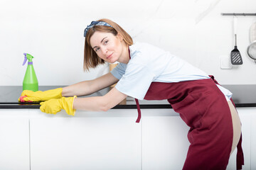 Young attractive housewife in red apron and yellow rubber gloves cleaning table with rag and bottle...