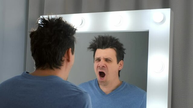 Tired sleepy man with a hangover looks in the mirror at his reflection yawns in morning. He rubs his swollen face and looks at his reflection in the mirror with light bulbs. Consequences of drinking.