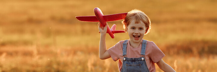 The girl runs across the summer field and launches a toy plane into the sky. A child plays aviator or pilot in a field at sunset, a happy childhood. Banner for website header design with copy space. © Kaplitskaya Love