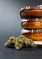 Dry buds of medical marijuana close-up, in the background, donuts covered with chocolate icing stand in a stack.  On a dark background