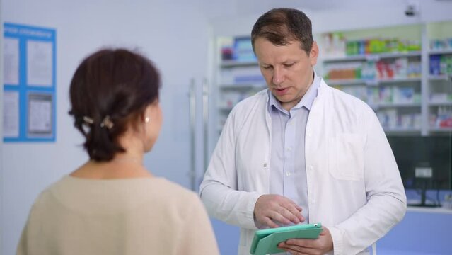 Portrait of serious man in uniform consulting woman standing in pharmacy indoors. Male Caucasian pharmacist talking in slow motion with female client in drugstore