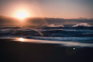 Rough ocean waves with spray crashing on volcanic black sand beach at dawn with low sun and lens...
