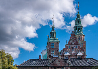 Copenhagen, Denmark - September 13, 2010: Rosenborg Slot NW red-stone facade top showing statues and 2 green steeples on towers under blue cloudscape and green foliage on side
