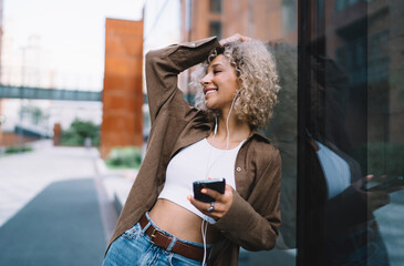 Cheerful ethnic woman listening to music from earphones and standing on street with smartphone