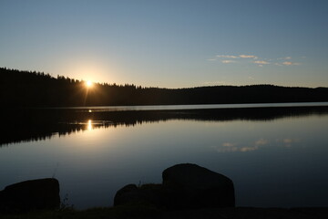 Obraz premium Calm lake with reflection of sun setting down behind forest