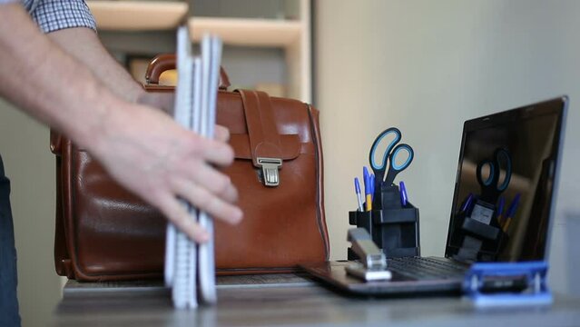 An unknown man takes documents from the table, puts them in a briefcase, and leaves work. End of the working day in the company.