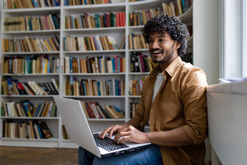 Young hispanic student studying inside academic library among bookshelves, man typing on laptop keyboard, smiling contentedly sitting near window.