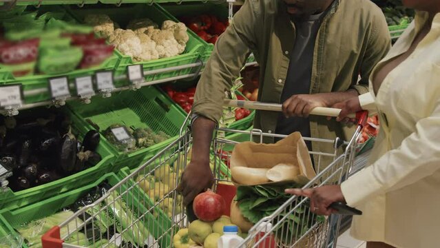 African American young couple putting fresh vegetables in trolley while doing shopping in supermarket