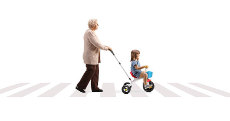 Full length profile shot of a grandmother pushing a child on a tricycle over pedestrian crossing