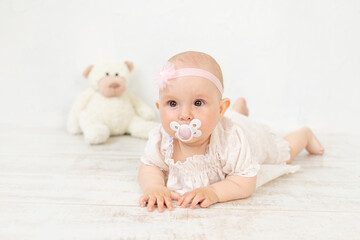 a baby girl with a pacifier in her mouth in a white dress with a toy lies on her stomach for six...
