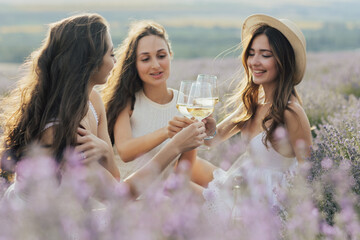 A beautiful happy young girlfriends clinking glasses with white wine at summer picnic in a lavender...