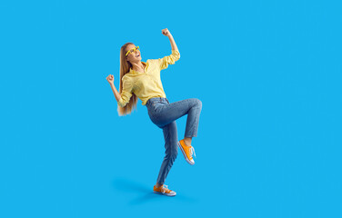 Happy emotional teen girl sincerely rejoices in her success isolated on light blue background. Stylish excited caucasian young woman in jeans, sneakers and shirt happily clenching fists. Full length.