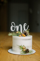 Vertical picture of white cake with sigh on top one for one year child’s birthday. Cáale is decorated with pine branches and pine corns on table.