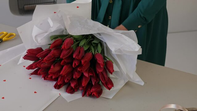 Caucasian lady florist collects a lush bouquet of tulips. A professional makes a fresh bouquet. Small business. A woman crumples paper (Crepe) and wraps a bouquet of tulips in it