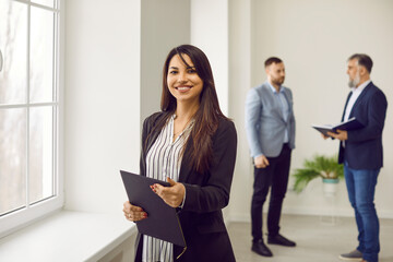 Portrait friendly female business consultant, secretary, estate agent, assistant manager at work. Happy beautiful smiling young woman with clipboard standing in office with other workers in background