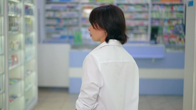 Back view Caucasian woman walking along rows of shelves in drugstore looking around. Tracking shot of thoughtful female customer choosing medications in pharmacy indoors. Slow motion