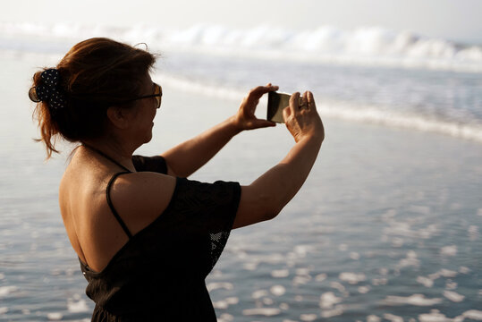 Woman on beach taking pictures of ocean waves with her phone's camera.