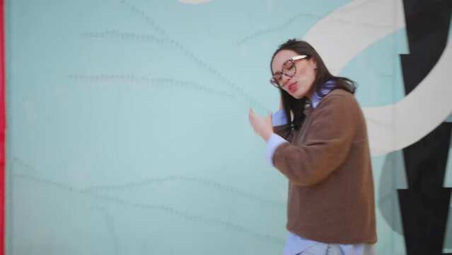 Happy woman in glasses standing outdoor near colorful wall smile and talking something. Positive female fix hair laught an tel a story 