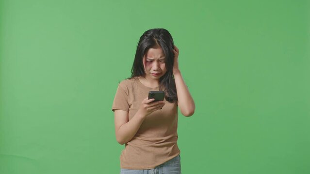 Young Asian Woman Victim Of Violence With Bruise On Face Looking At Smartphone And Crying While Standing On Green Screen Background In The Studio
