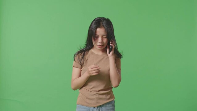 Young Asian Woman Victim Of Violence With Bruise On Face Crying And Talking On Smartphone Asking For Help On Green Screen Background In The Studio
